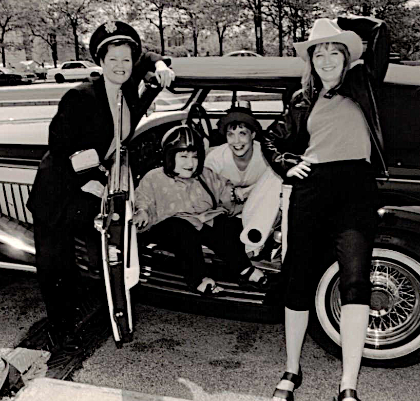 A black and white photo of four women wearing different hats: Tekki Lomnicki (motorcycle helmet); Judith Harding (Irish cap); Lotti Pharriss Knowles (cowboy hat) standing or sitting in a vintage convertible.